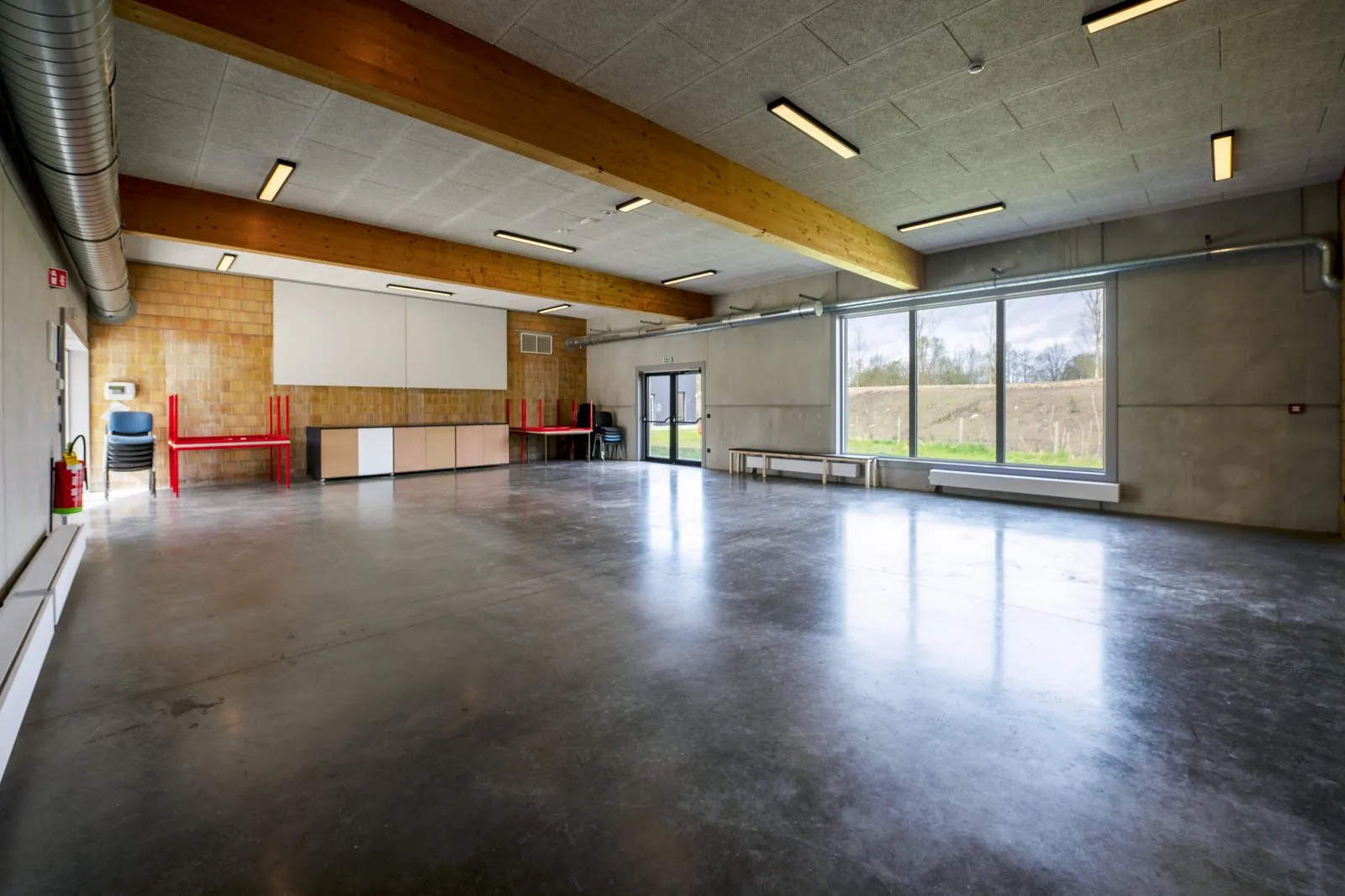 De grote zaal met houten spanten, een projectiescherm, opgestapelde stoelen en een grote raampartij op het groen