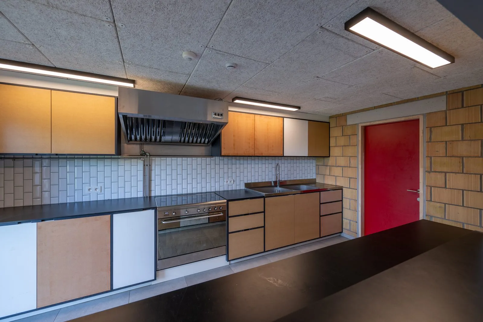 De keuken met inox werkbladen, een grote dampkap en een rode deur