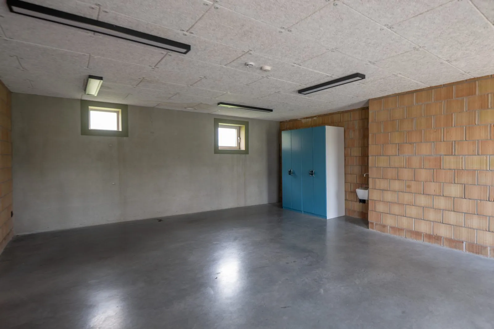 Een leeg lokaal met een betonvloer, hoge vensters, een bakstenen muur en een blauwe kast in de hoek